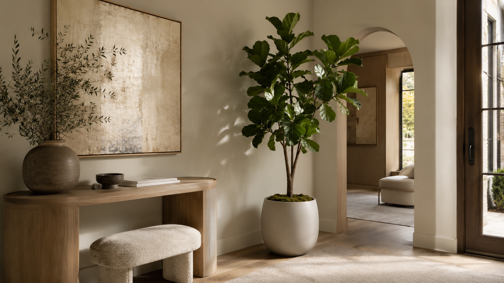Horizontal view of a refined entryway with a faux fiddle leaf fig, arched doorway, wood console, and soft natural light.