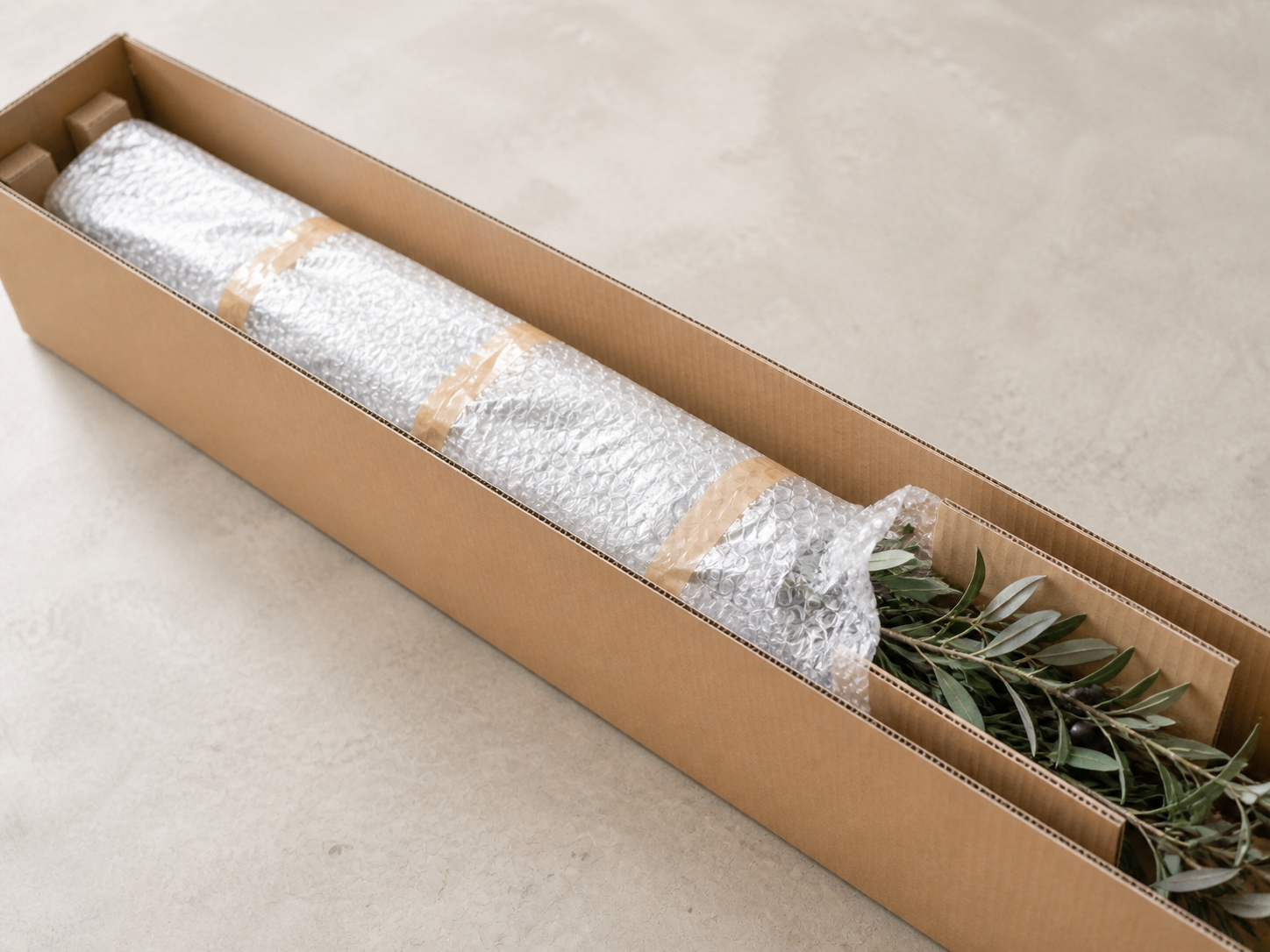 Long cardboard box with a faux olive tree wrapped in bubble wrap for protective shipping.